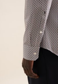 Camicia a maniche lunghe di colore bianco con un motivo geometrico rosso e blu. Presenta polsini con bottoni e una vestibilità rilassata. Realizzata in tessuto leggero.