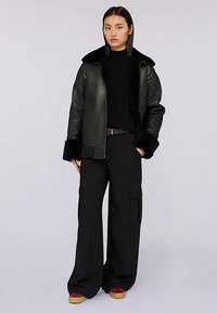 Svart läderbomberjacka med pälskrage, långa svarta cargobyxor och klumpiga skor med vinröd accent. Slät textur, modern design.