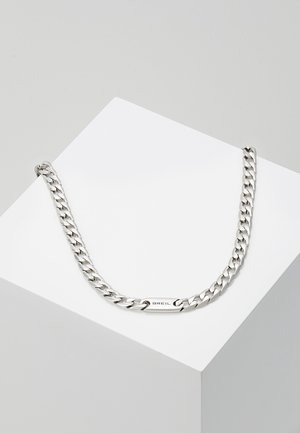 Collier chaîne en argent avec des maillons plats entrelacés et une plaque rectangulaire gravée de "BREIL" posée sur une surface géométrique blanche.