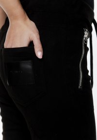 Czarne zamszowe spodnie posiadają boczną kieszeń z srebrnym zamkiem błyskawicznym oraz teksturowaną skórzaną łatę z nazwą marki.
