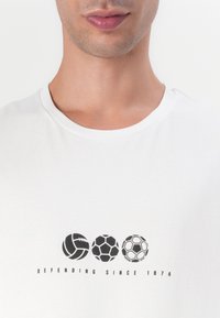 Vit kortärmad t-shirt med tre svarta sportbollsgrafik och texten "FÖRSVARAR SEDAN 1874" under dem. Slät bomullstyg.