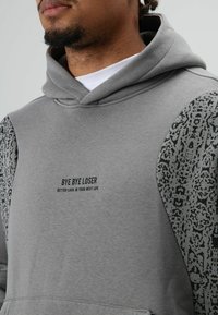 Man met een grijze hoodie met patroonmouwen en de tekst "BYE BYE LOSER, BETTER LUCK IN YOUR NEXT LIFE" op de borst.