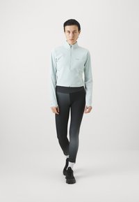 Lyseblå zip-up jakke med lange ermer, paret med leggings i svart til grå gradient. Modellen har svarte sko med hvite sokker.
