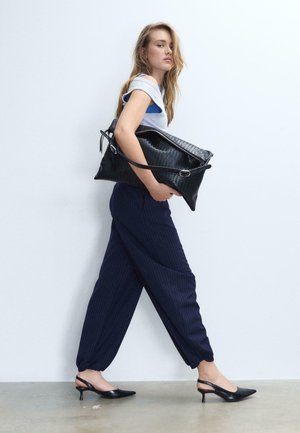 Ženska hodi v mornarsko modrih hlačah z ozkimi črtami in črnih salonarjih, pod roko nosi veliko teksturno črno torbico na belem ozadju.