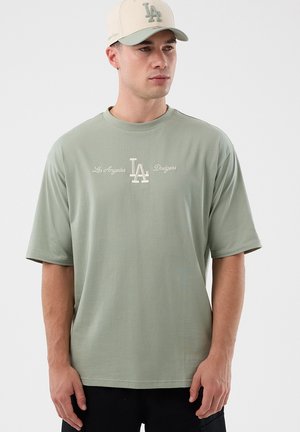 Mężczyzna w jasnobrzozowej koszulce Los Angeles Dodgers i kremowej czapce bejsbolowej z logo LA, stojący na jednolitym tle.
