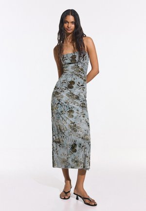 Femeie cu păr lung și închis la culoare, purtând o rochie florală fără bretele, până la glezne, și sandale negre cu toc, stând în fața unui fundal alb simplu.