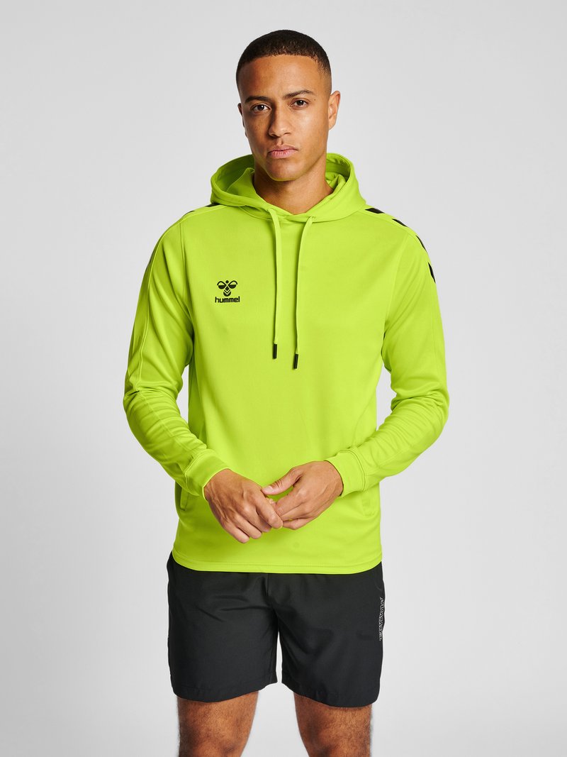 Mann, der einen neongrünen hummel-Hoodie und schwarze Shorts trägt, steht mit verschränkten Händen vor einem grauen Hintergrund.