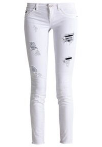 Jean blanc extensible avec des patchs usés, comprenant des trous effilochés et des détails de couture variés, présentant une coupe slim et un design classique à cinq poches.