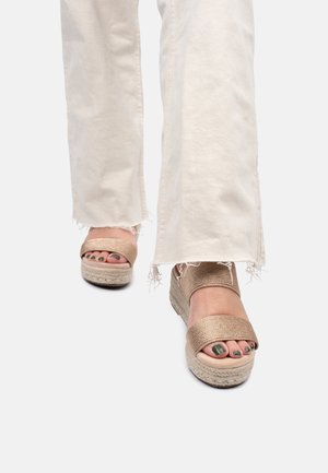 Person iført hvide bukser med frynset kant og beige vævede platformsandaler med mørkegrøn neglelak på tæerne.