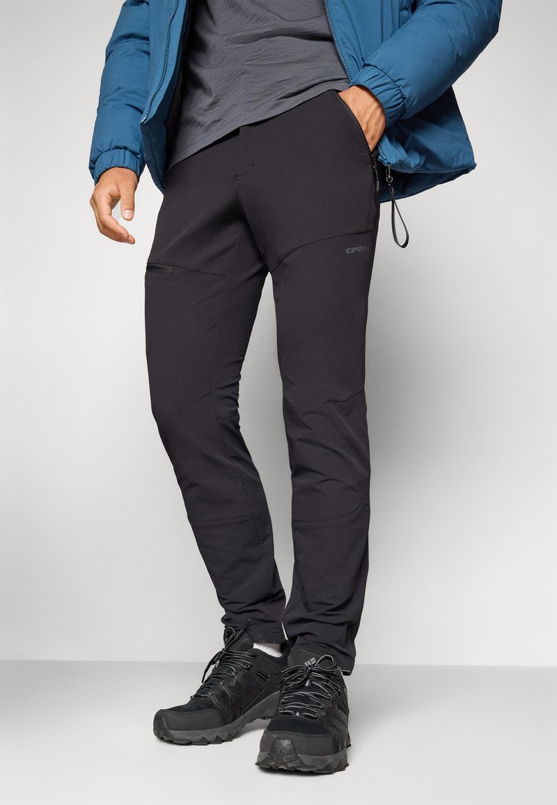 Schwarze Outdoor-Hosen aus Stretchstoff, mit Reißverschlusstaschen, schlanker Passform und verstärkten Knien für Langlebigkeit. Kombiniert mit schwarzen Wanderschuhen.