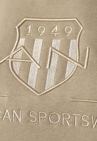 La tela beis presenta un escudo bordado con rayas verticales, el año "1949" y el texto "ROPA DEPORTIVA PARA HOMBRE" en un sutil hilo color crema.