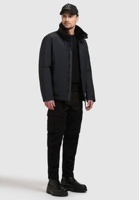 Mężczyzna w czapce baseballowej, czarnej kurtce zapinanej na zamek z futrzanym kołnierzem, czarnych spodniach cargo i czarnych butach na sznurowadła stoi na białym tle.