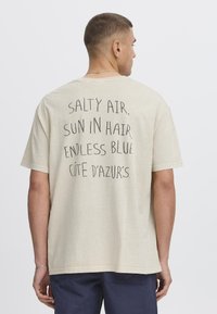 Beige cotton t-shirt featuring a relaxed fit, short sleeves, and text on the back: "SALTY AIR, SUN IN HAIR, ENDLESS BLUE CÔTE D'AZUR'S."