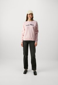 Felpa rosa chiaro con ricamo navy "Tommy Hilfiger", abbinata a jeans grigio scuro e scarpe nere, completata con un cappellino beige.