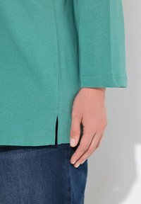 Türkises Langarmhemd mit seitlichen Schlitzen, aus weichem Baumwollstoff gefertigt. Kombiniert mit dunklen Jeans, die den Kontrast in Farbe und Textur hervorheben.