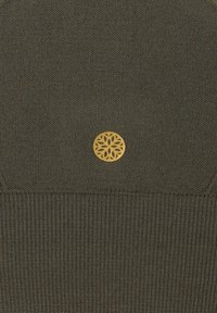 Dunkelgrüner Strickstoff mit gerippter Textur, der ein goldfarbenes, rundes Logo mit einem komplizierten Muster zeigt, das sich nahe der Oberseite befindet.