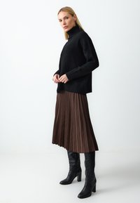 Maglione nero a collo alto con polsini a coste, abbinato a una gonna midi plissettata marrone e stivali neri con tacco al ginocchio. Silhouette semplice ed elegante.
