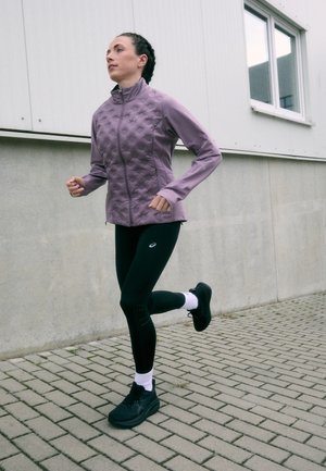 Ljubičasta teksturirana trkačka jakna s visokim ovratnikom, uparena s crnim tajicama koje imaju suptilne perforirane detalje. Crne trkačke tenisice i bijele čarape.