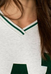 Szary bawełniany T-shirt z zielono-białym paskowanym dekoltem w kształcie litery V oraz dużym zielonym napisem na przodzie. Miękka faktura, casualowy design.