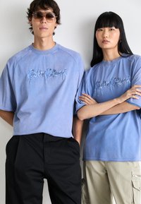 Ljusblå bomullströjor med broderad text i droppande stil. Modellens svarta byxor och beiga cargobyxor fulländar looken.