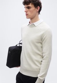 Jeune homme portant un pull crème superposé à une chemise blanche à col, tenant une mallette noire rectangulaire sur un fond clair uni.