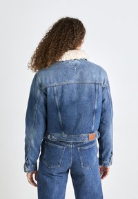 Chaqueta de mezclilla con un lavado azul claro, cuello de borrego y costuras visibles. Cuenta con bolsillos frontales y detalles de parche de cuero en la parte posterior.