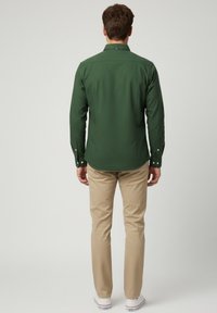 Camisa de manga larga verde oscuro, con cuello y puños abotonados, combinada con pantalones beige y zapatillas blancas. Tejido suave con un corte ajustado.