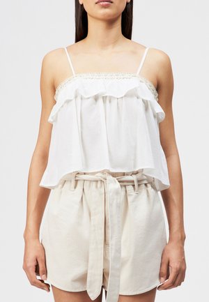 Kvinde iført hvid, ærmeløs, flæset top og beige højtaljede shorts med stofbælte bundet i en sløjfe ved taljen.