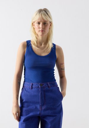 Ung kvinde med blondt hår iført en blå tanktop og blå højtaljede jeans, stående med den ene hånd i lommen mod en hvid baggrund.