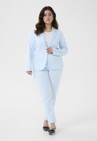 Světlomodrý oblek s na míru šitým blejzrem a slim fit kalhotami. Hladká textura, vykrojené klopy a minimální detaily. Nosené s černými balerínami.
