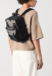 Sac à dos noir et gris avec des bretelles réglables, des détails en cordon élastique, une poche frontale et un tissu texturé, porté par-dessus un pull beige.