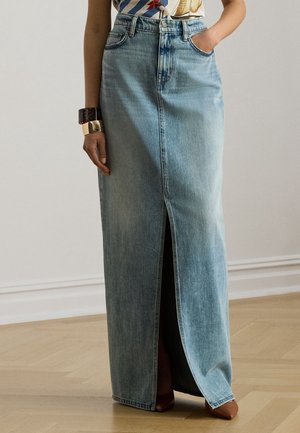 Femme portant une jupe longue en denim bleu clair taille haute avec une fente à l'avant, associée à des chaussures pointues marron et des bracelets au poignet gauche.