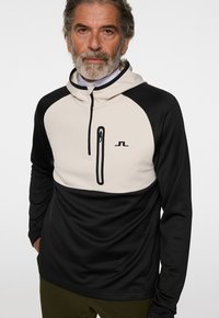 Pull à capuche en tissu doux, avec une partie inférieure noire et une partie supérieure crème, doté d'une poche zippée sur la poitrine et d'un logo.