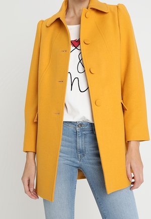 Femme portant un manteau jaune moutarde boutonné sur un t-shirt blanc à motifs et un jean bleu clair, debout devant un fond uni.