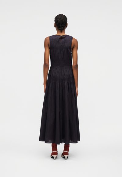 Femme aux cheveux tressés portant une robe noire sans manches jusqu'aux chevilles et des talons blancs avec des chaussettes couleur rouille, debout de dos.