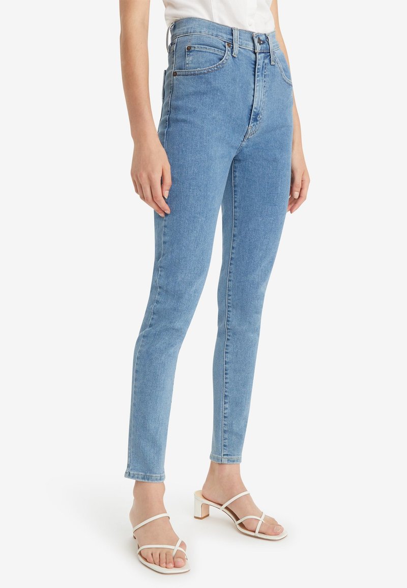 Jean en denim bleu clair taille haute, avec une coupe slim. Présente un design classique à cinq poches et des coutures discrètes. Associé à des sandales blanches.