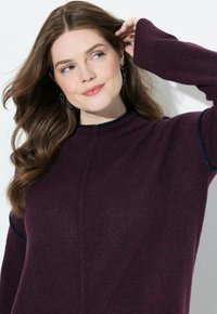 Donna con lunghi capelli castani, che indossa un maglione viola e orecchini a cerchio, toccandosi i capelli, con uno sguardo di lato e un leggero sorriso.