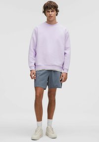 Sweatshirt lilas avec des poignets côtelés, associé à un short gris clair. Des baskets blanches et des chaussettes blanches complètent la tenue. Style et coupe décontractés.