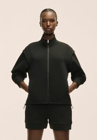 Sudadera negra con cremallera, hecha de un tejido suave, con cuello alto, mangas relajadas y bolsillos laterales, combinada con pantalones cortos a juego.