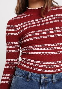 Rode gebreide trui met witte zigzagstrepen, een hoge hals en een structuurpatroon. Gecombineerd met blauwe denim hoge taille jeans.
