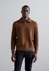 Homme se tenant contre un fond uni, portant un pull zippé marron et un pantalon sombre, regardant vers l'avant avec une expression neutre.