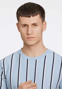 Ljusblå randig t-shirt med mörk marinblå och vita vertikala linjer, rund halsringning och en strukturerad tyg. Modell som visar avslappnad pose.