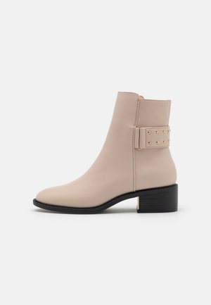 Botín beige hasta el tobillo con un acabado de cuero suave, punta redondeada y tacón cuadrado. Presenta una correa lateral decorada con tachuelas doradas.