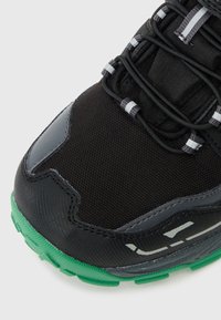 Fekete sportcipő, textúrált hálós felsőrésszel, szürke kiegészítőkkel, fényvisszaverő részletekkel és zöld gumitalppal, amely tapadási mintákkal rendelkezik.