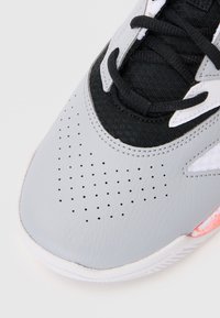 Grey athletic shoe with a textured leather upper, black mesh accents, circular perforations, white sole, and a pink detail at the heel.