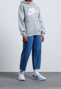 Grå Nike-hoodie med en vit logo, parat med lösa blå denimjeans och vita sneakers med grå och marinblå accenter.
