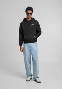 Svart bomullshoodie med frontlomme, hette og trykket tekst. Kombinert med lyseblå vide jeans og hvite sneakers.