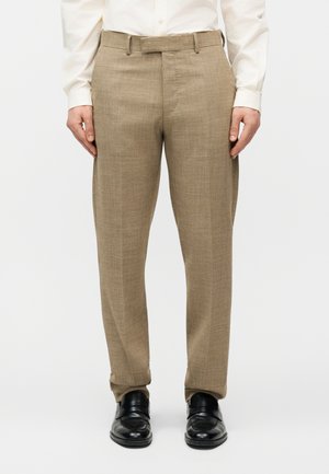 Mand iført beige bukser med lige pasform, hvid skjorte og sorte lædersko, stående mod en ensfarvet hvid baggrund.