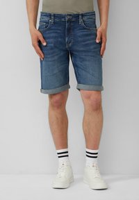 Shorts en denim bleu foncé avec des ourlets retroussés, cinq poches et une fermeture éclair, portés avec des baskets blanches et des chaussettes rayées.