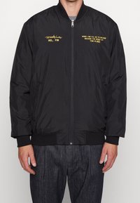 Giacca bomber nera in materiale sintetico con chiusura a zip, polsini a costine e testo ricamato giallo ben in vista sul petto.